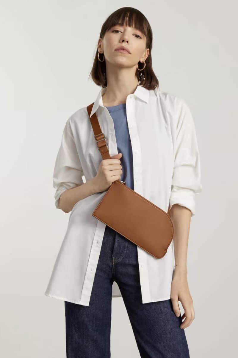 Woman wears honey toned sling bag from Everlane.