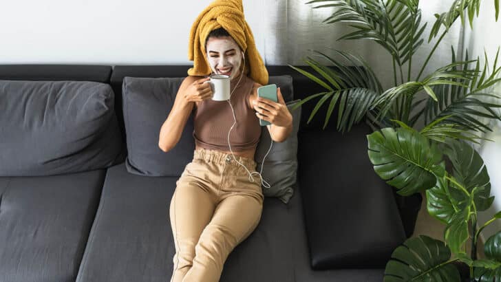 Woman wearing DIY beauty mask at home sips coffee and smiles at her smartphone.