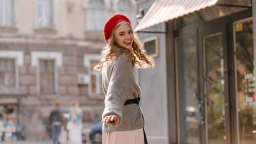 Woman wearing baggy sweater and hat smiles in street.