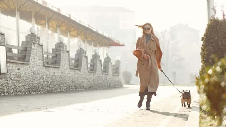 Woman wearing trench coat walks her dog outside.