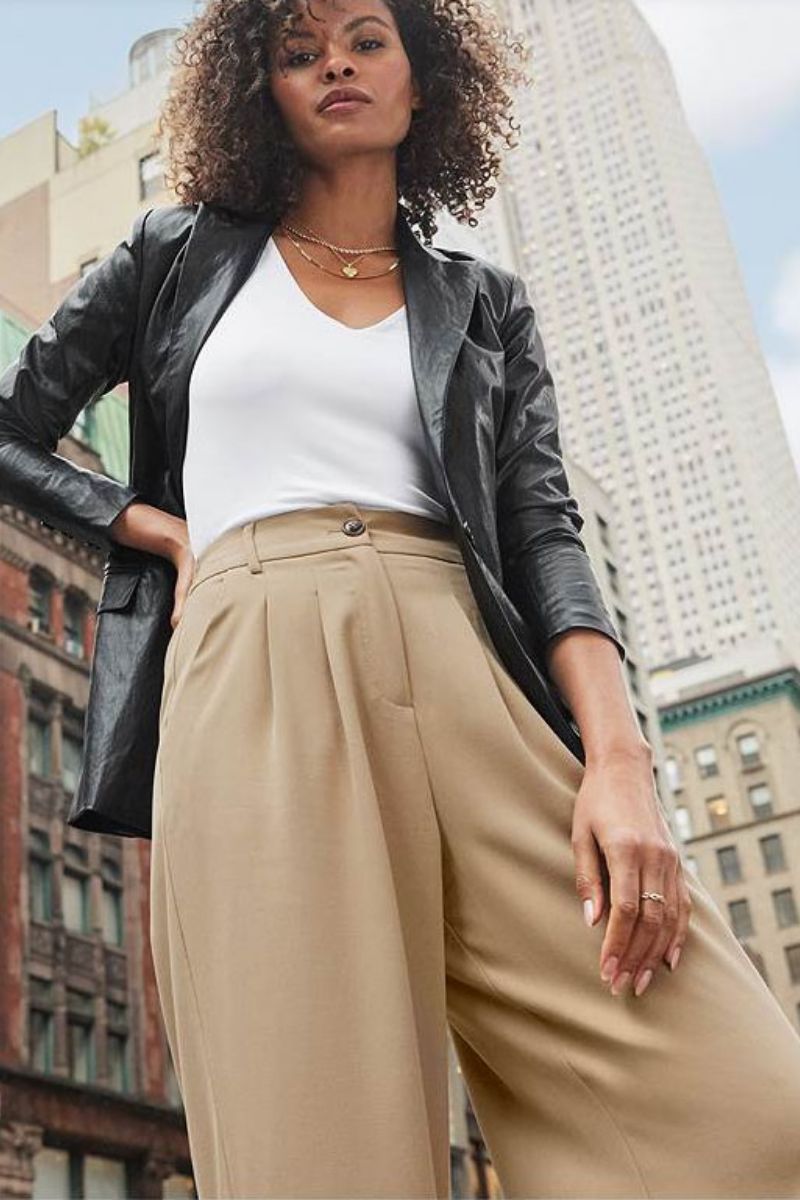 Model looking down at camera wears leather blazer, white top, and beige trousers.