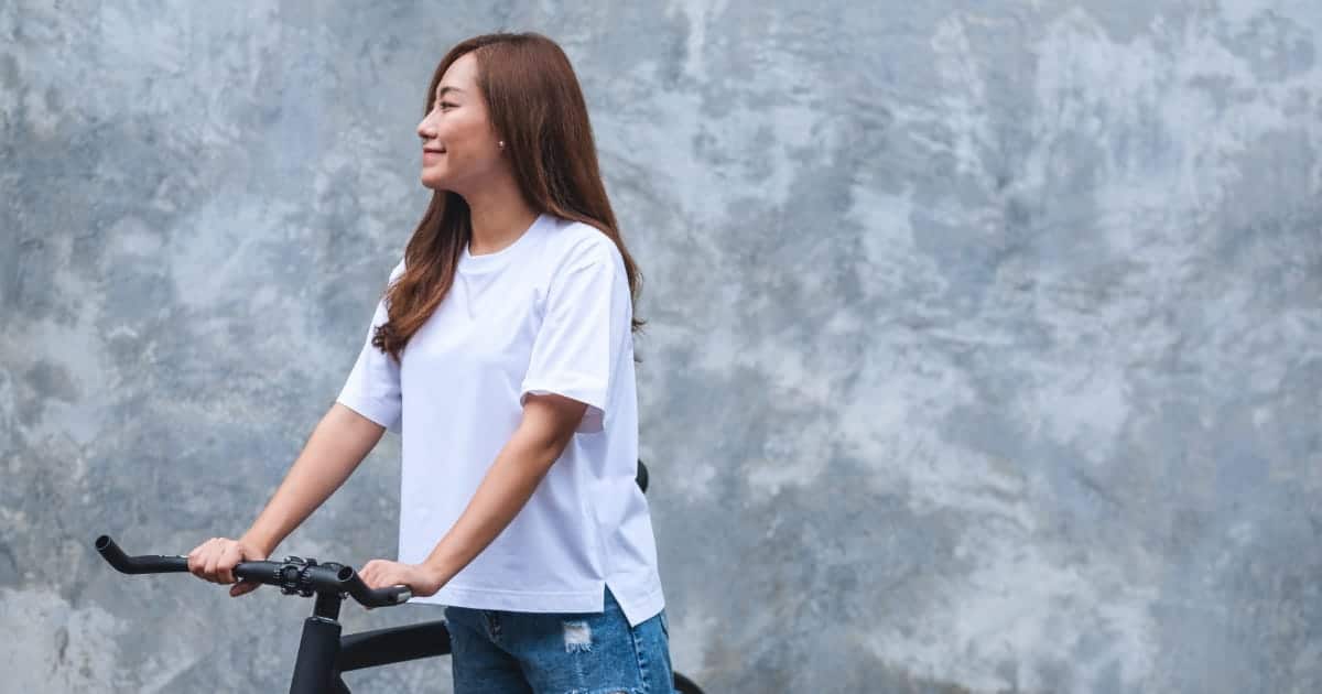 Woman wearing white tee shirt standing outside with bicycle.
