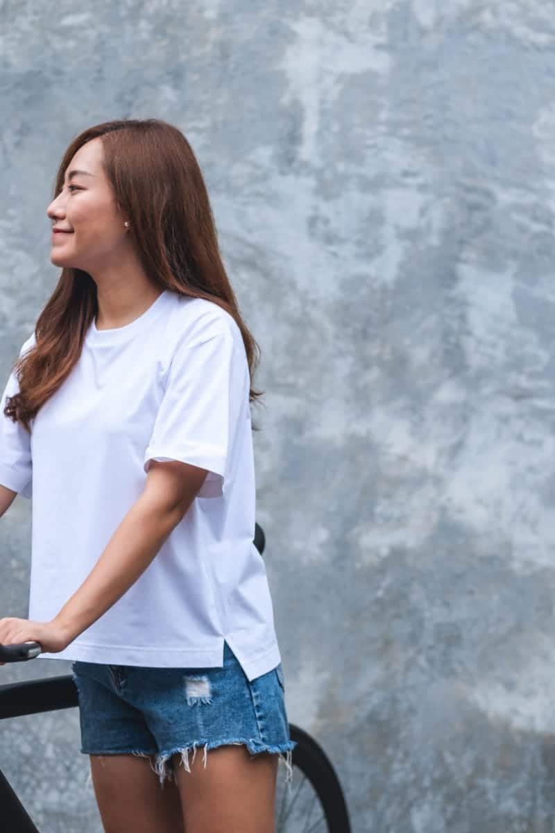 8 White Tees to Add to Your Wardrobe 1 Woman wearing white tee shirt standing outside with bicycle.