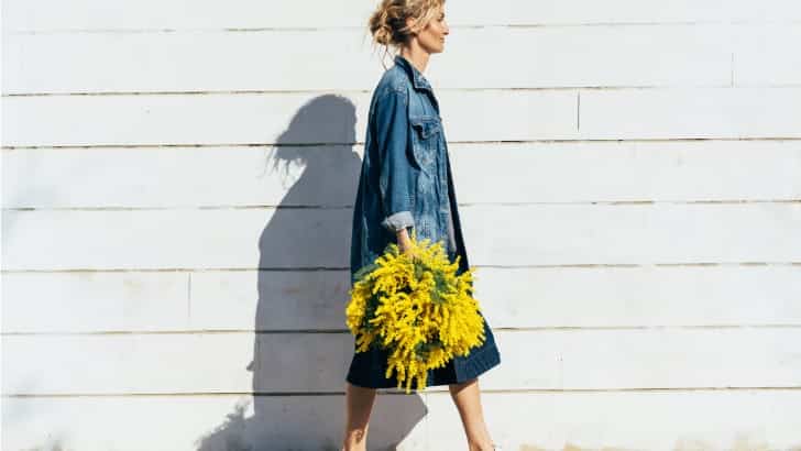 Woman wearing fall denim pieces walks in front of wall holding flowers.