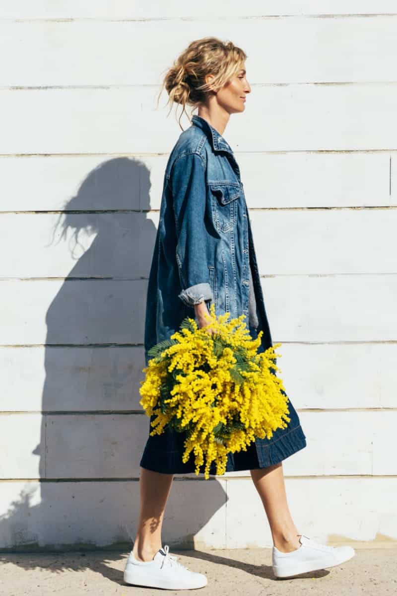 6 budget-friendly fall denim pieces you'll love 1 Woman wearing fall denim pieces walks in front of wall holding flowers.