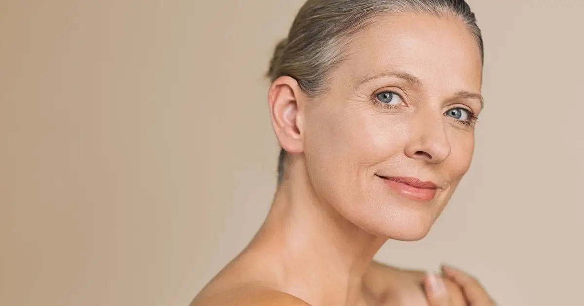 Smiling mature woman with hair pulled back in front of beige background.