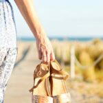 View from back of woman holding trendy spring sandals at beach.