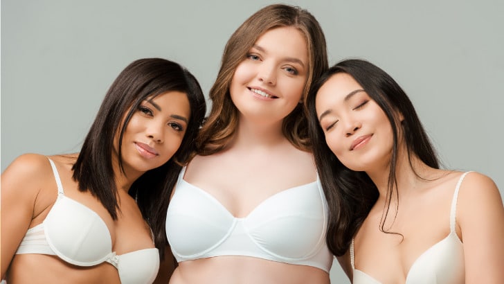 Three smiling women wearing bras in front of gray background.