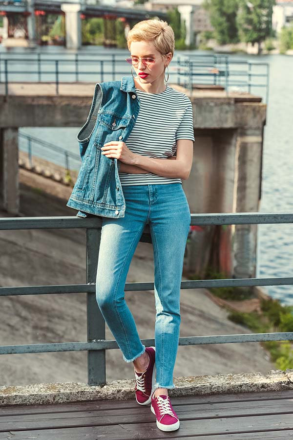 Short-haired woman wearing glasses and blue jeans leans against metal fence outside.