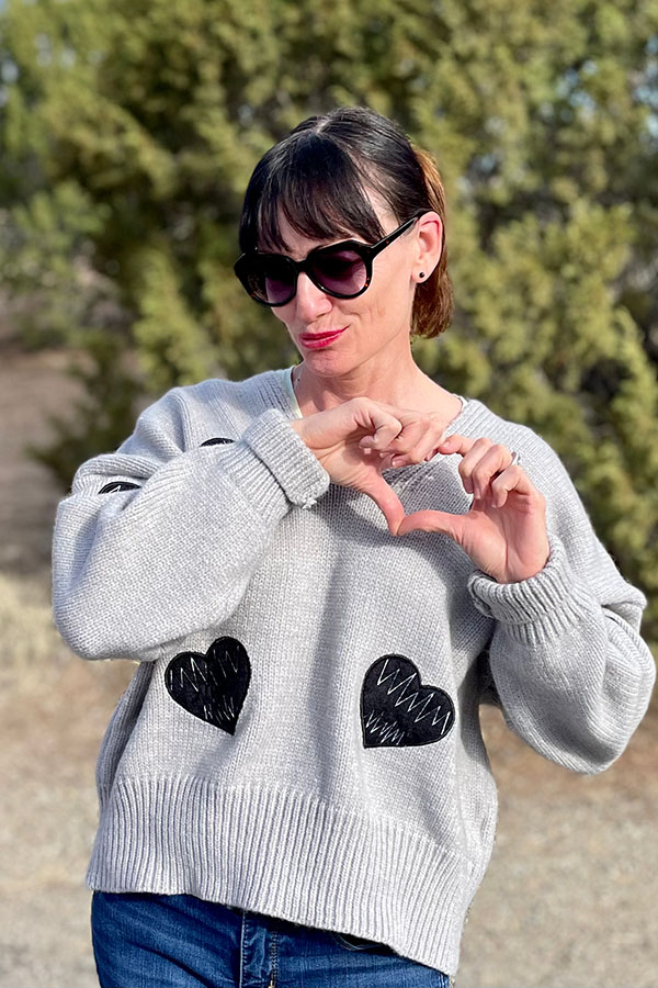 Catherine Brock wears gray sweater with black heart detail from Chicwish, while making a heart-shape with her fingers.