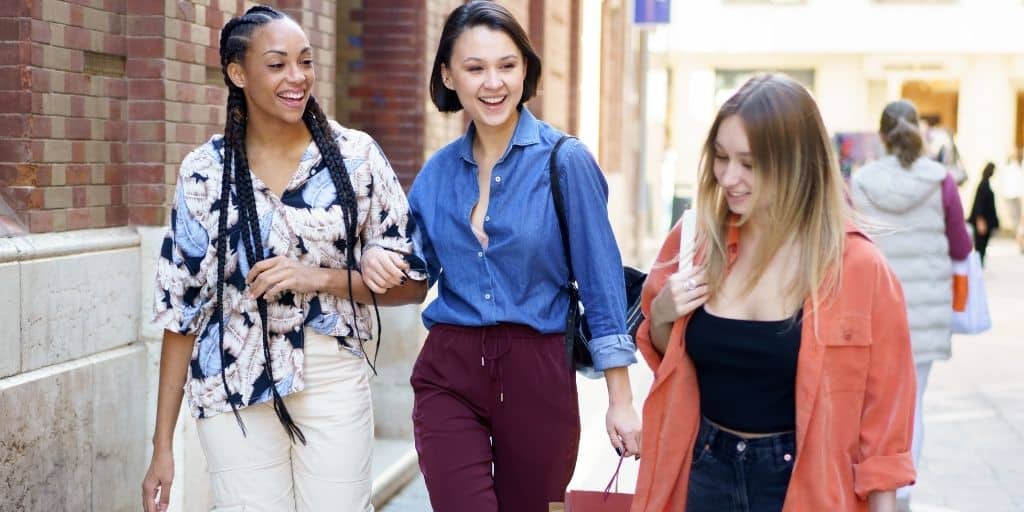 Three friends laugh together while walking on street with shopping bags.