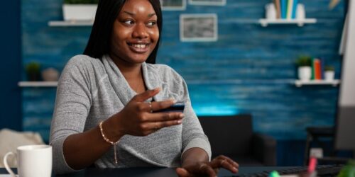 Smiling person holds phone while looking at laptop.