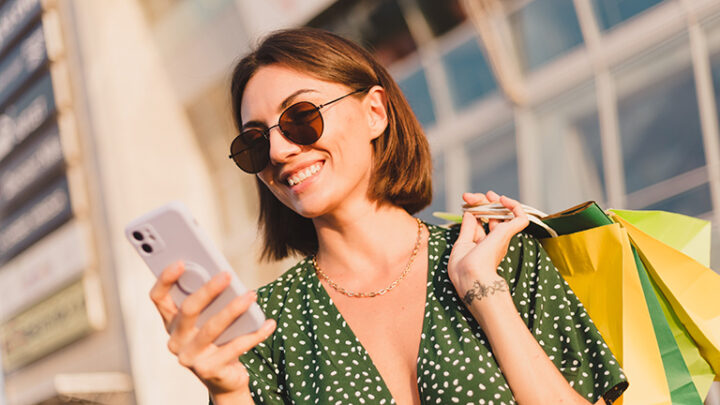 Smiling, stylish shopper stands outside holding smartphone.