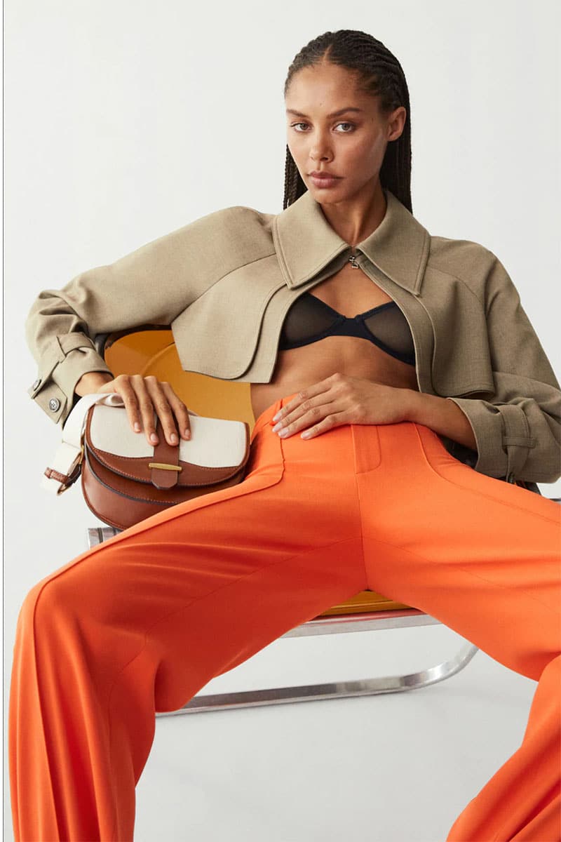 Modelka w pomarańczowych spodniach podczas siadania trzyma w pasie torebkę crossbody.