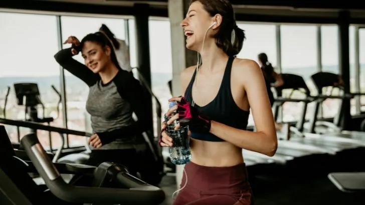 Two women in the gym laugh while wearing workout clothes.