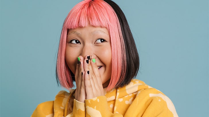 Woman with pink hair, cut in a bob style smiles with hands over mouth.