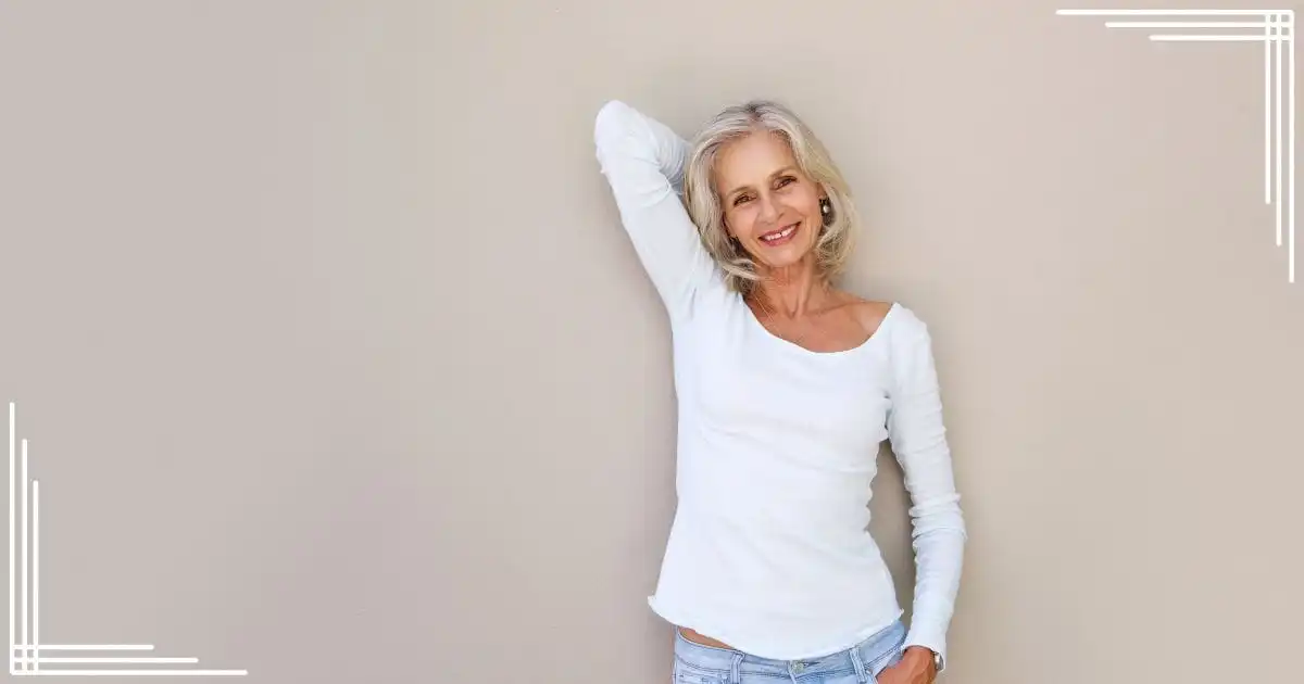 Older woman wearing white long-sleeved t-shirt smiles in front of beige background.