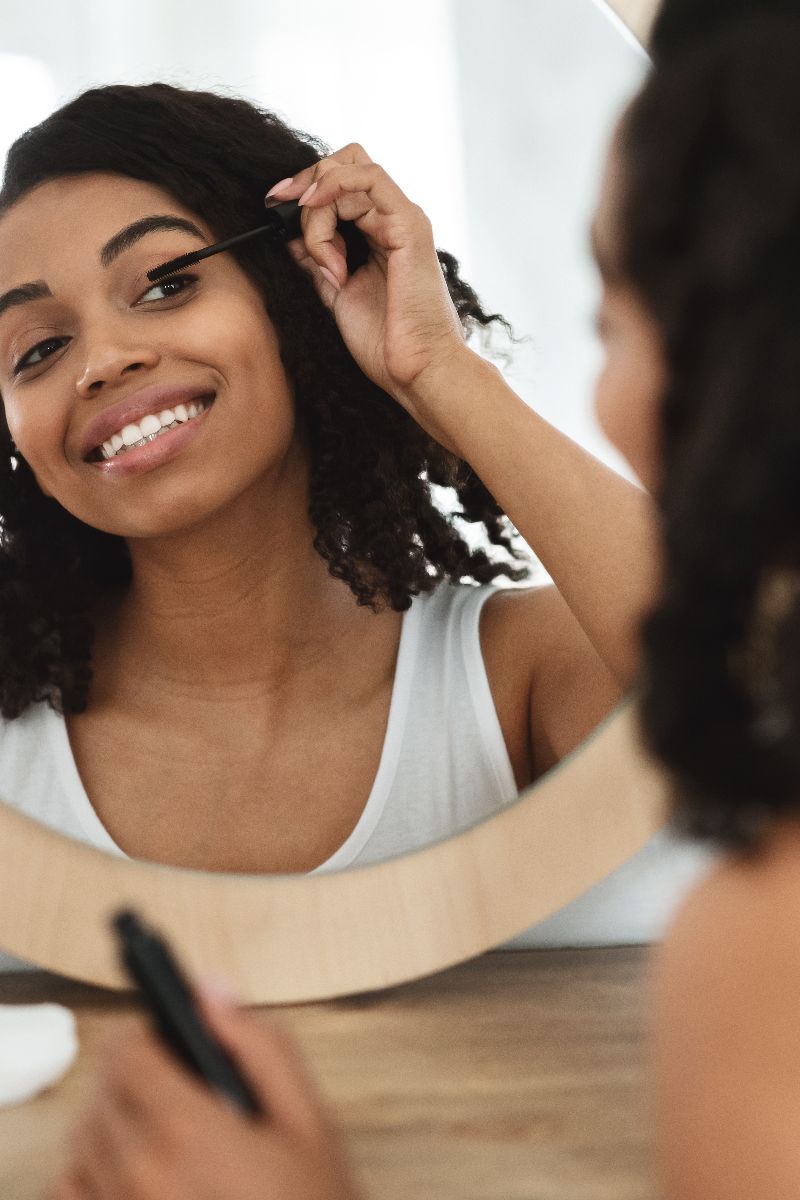 Woman applies mascara while looking in mirror.